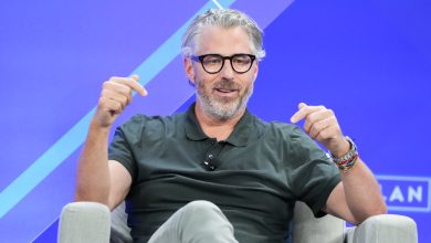 Casey Wasserman wears black glasses and a polo shirt while sitting on stage during a panel discussion