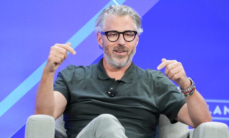 Casey Wasserman wears black glasses and a polo shirt while sitting on stage during a panel discussion