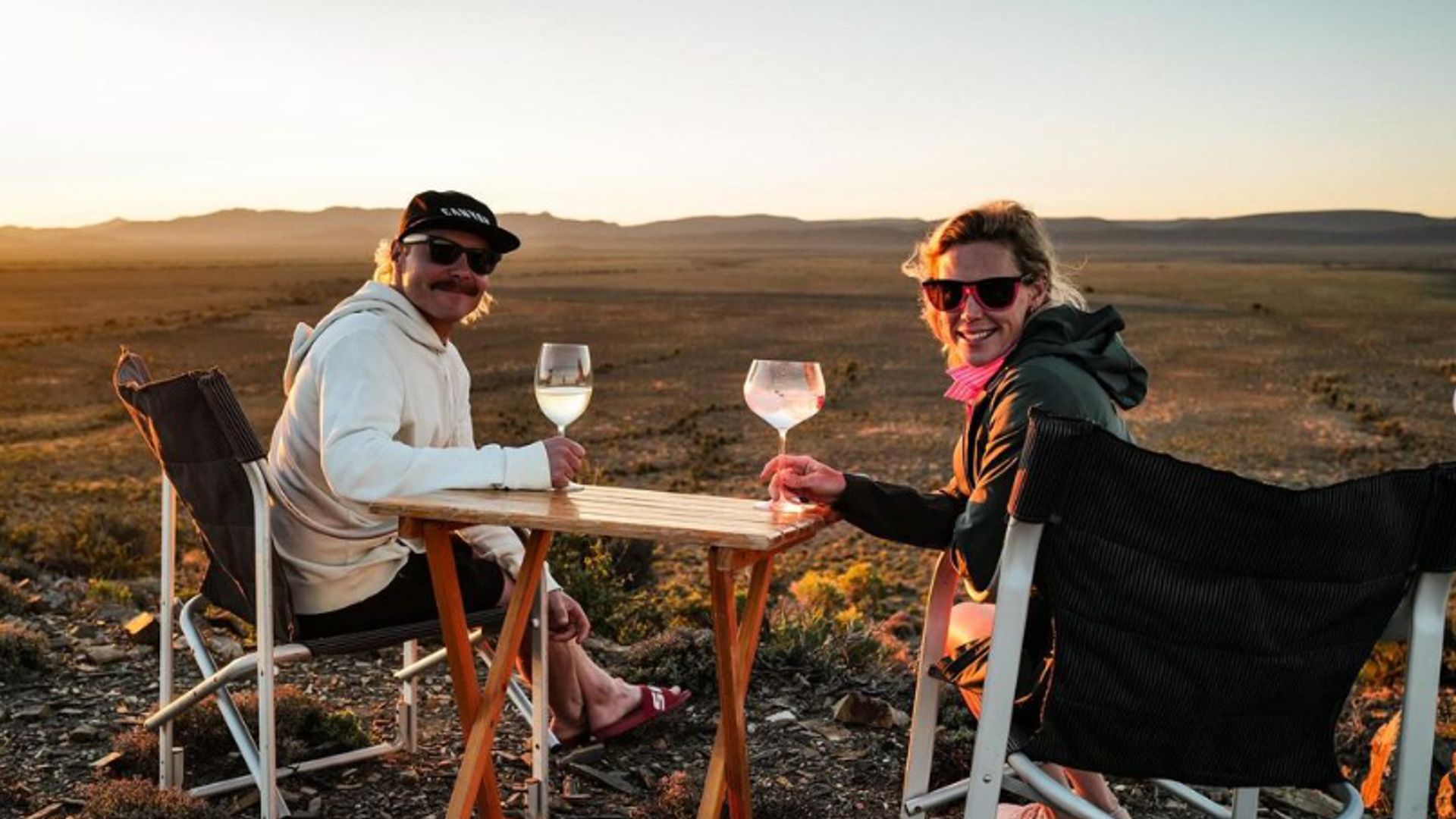 Valterri Bottas and Tiffany Cromwell with glasses of alcohol sat at a wooden table outfit