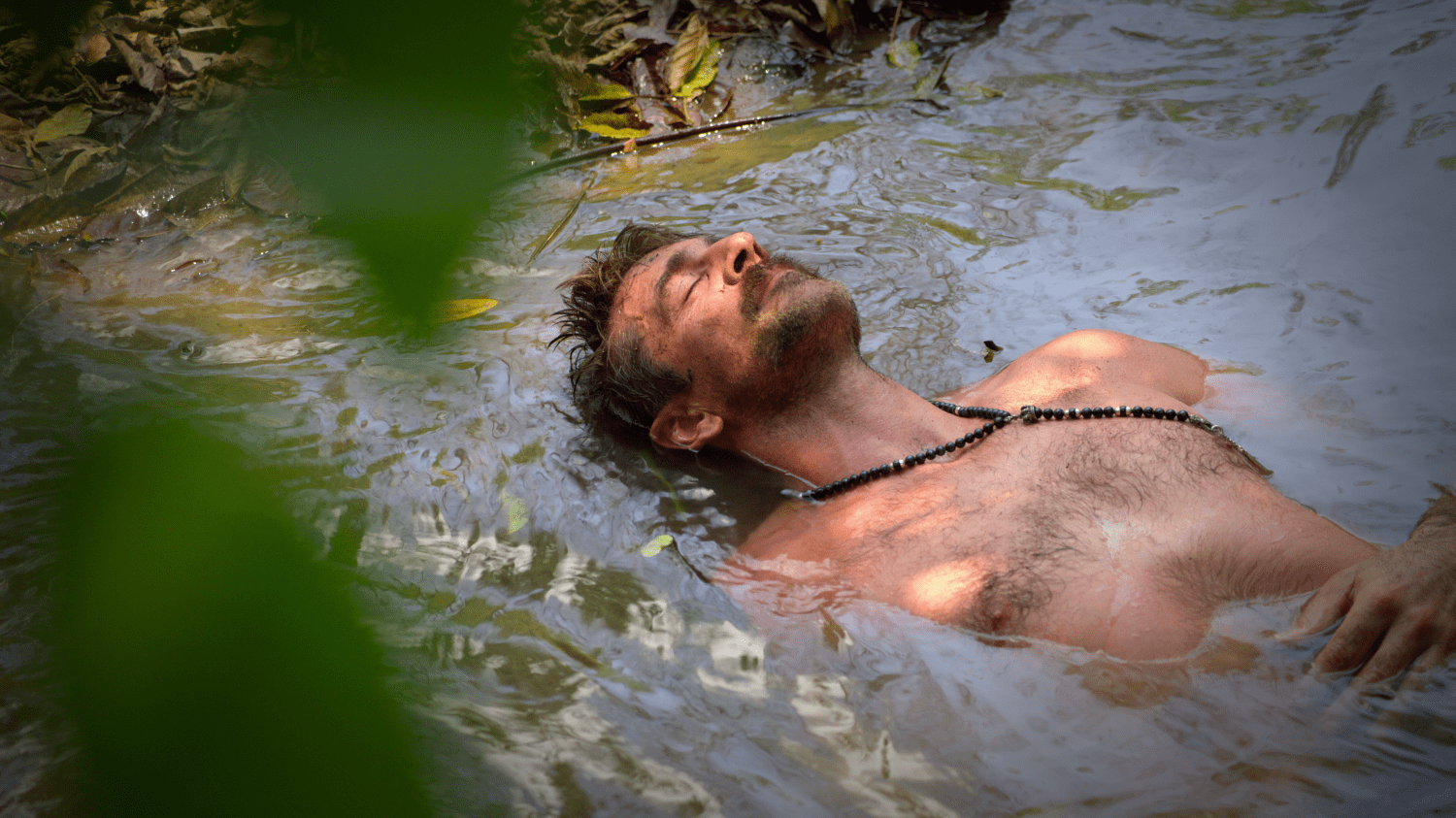 Aaron Rodgers lays in a muddy pool of water with his eyes closed while shirtless and wearing black beads.