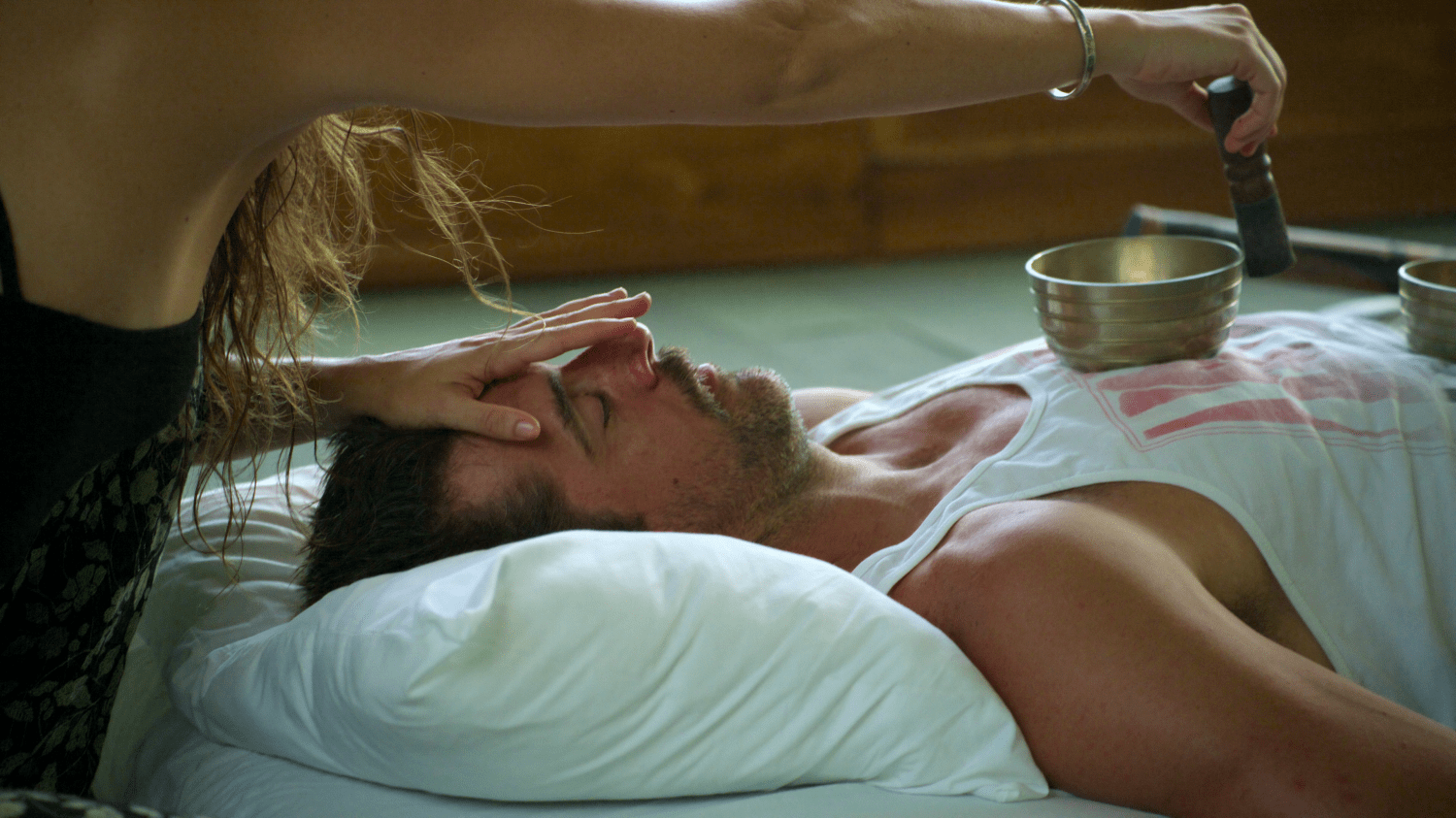 Aaron Rodgers lays on the ground with his eyes closed while a woman rubs a golden bowl with a wooden tool for a sound bath.