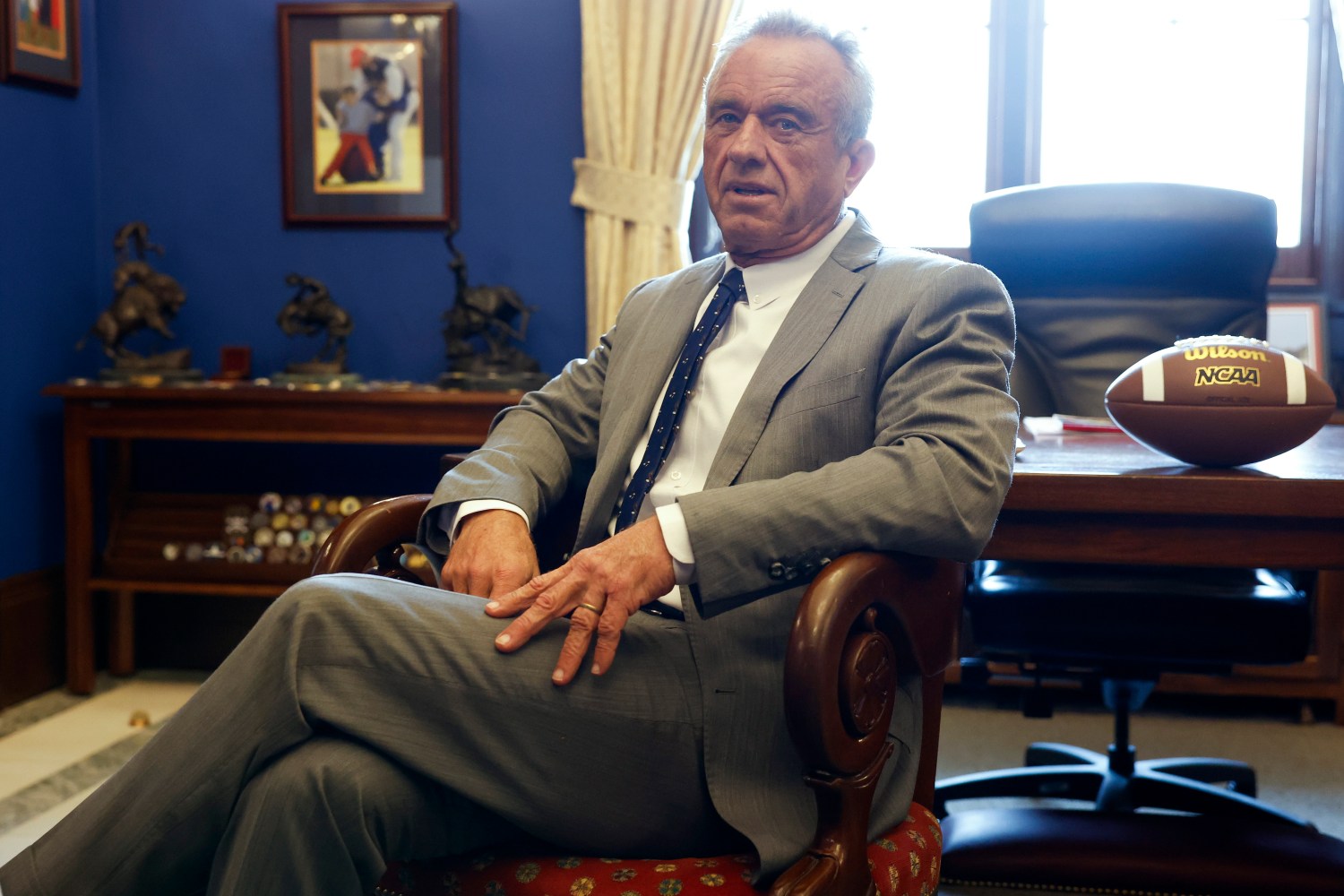 Robert F. Kennedy Jr. sits in an office leaning back in a chair with a table behind him that is holding a Wilson NCAA football.