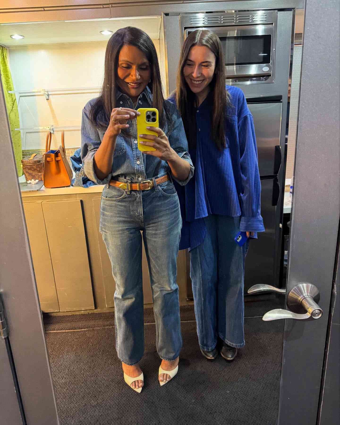 Mindy Kaling wearing denim standing next to a friend in kitchen mirror