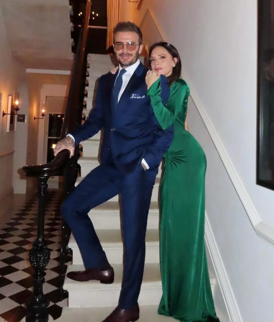 Victoria Beckham in a green floor length dress and David Beckham in a blue suit standing on their stairs in the hallway