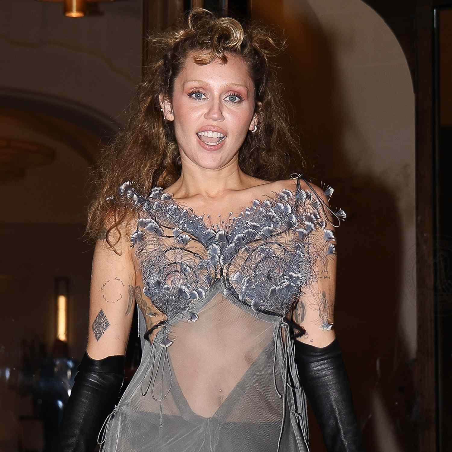 Miley Cyrus poses in a striking sheer dress with feathers outside her hotel, completing the look with black gloves.