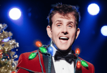 Joey McIntyre wears a red velvet blazer with bright Christmas bulbs draped across his torso while smiling in front of a Christmas tree.