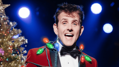 Joey McIntyre wears a red velvet blazer with bright Christmas bulbs draped across his torso while smiling in front of a Christmas tree.