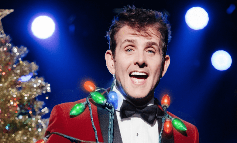 Joey McIntyre wears a red velvet blazer with bright Christmas bulbs draped across his torso while smiling in front of a Christmas tree.