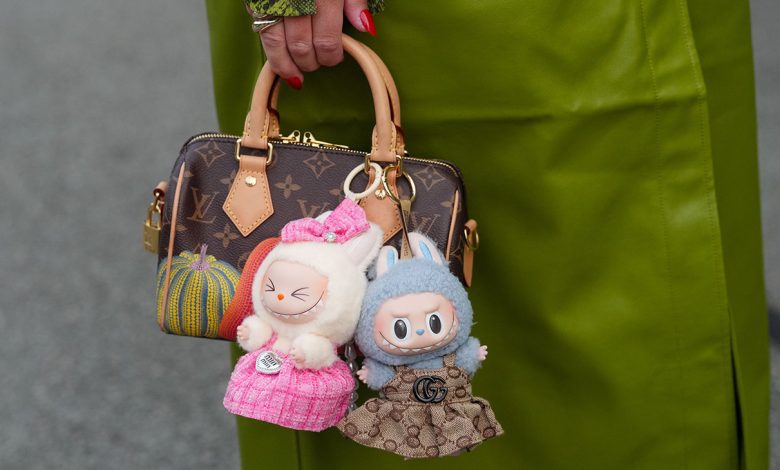 A guest wears a bright olive-green leather coat with a green snake-print cuff peeking from the sleeve, a silver-toned ring, and a glossy red pointed manicure, while a brown Louis Vuitton Monogram coated-canvas mini top-handle bag with tan vachetta leather rolled handles, leather tabs, a gold-tone zipper and padlock, and a yellow-and-orange pumpkin motif is carried; two Labubu plush bag charms are attached-one cream plush figure in a pink tweed dress with a matching bow, the other blue-gray plush with bunny ears in a brown Gucci GG monogram dress finished with a black Double G emblem and gold-tone hardware attachments, outside Rotate, during Copenhagen Fashion Week, on August 07, 2025 in Copenhagen, Denmark